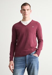 Pulover burgund cu decolteu în V, dintr-un material moale; design ajustat cu manșete și tiv ribbat. Purtați peste un tricou alb, asortat cu blugi albaștri.