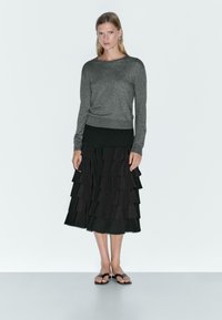 Un haut gris à manches longues associé à une jupe noire à volants multiples, portée avec des tongs noires. Tissu lisse avec un design superposé.
