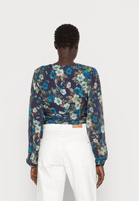 Blouse à manches longues à fleurs sombres avec manches froncées, ornée de fleurs bleues et crème sur fond bleu marine, portée avec un pantalon blanc.