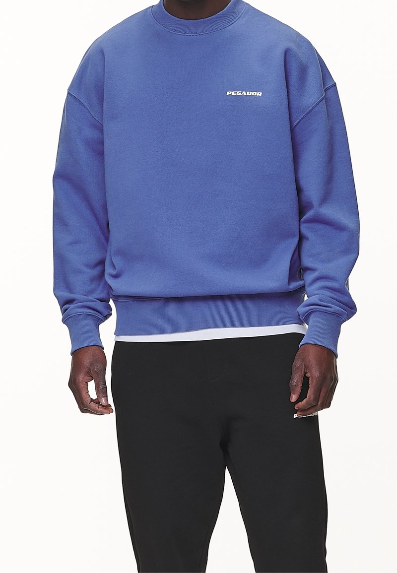 Un maglione a girocollo viola realizzato in misto cotone, con polsini e orlo a costine. Logo "PEGADOR" in bianco sul petto. Abbinato a pantaloni neri.