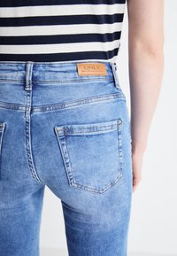Jeans de mezclilla de color azul claro con cintura alta, bolsillos traseros y un parche de cuero de la marca. La tela presenta una textura desgastada y sutiles detalles de costura.