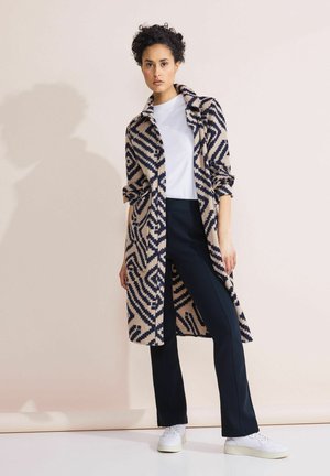 Cappotto lungo a motivi in beige con design geometrici blu navy, con colletto e maniche arrotolate, abbinato a pantaloni scuri e sneakers bianche.