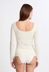 Donna che indossa un top crema a maniche lunghe con schiena scoperta e biancheria intima abbinata, in piedi davanti a uno sfondo bianco uniforme.
