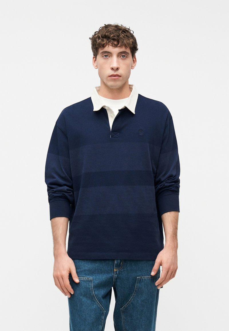 Νεαρός άνδρας φορώντας μπλε ναυτικό ράγκμπι μπλουζάκι με λευκό γιακά και μπλε τζιν, στέκεται μπροστά σε ένα απλό λευκό φόντο.