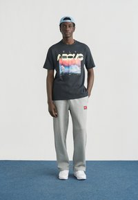 Μαύρο μπλουζάκι με πολύχρωμο σχέδιο σύννεφων, συνδυασμένο με γκρι φόρμα. Το μοντέλο φοράει λευκά αθλητικά παπούτσια και ανοιχτό μπλε καπέλο.