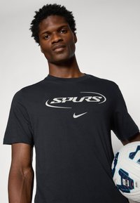 Sort T-shirt lavet af bomuld med korte ærmer, der har et stort gråt "SPURS" logo og en Nike swoosh. Modellen holder en fodbold.