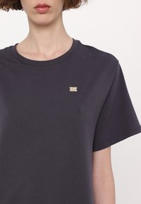 Tricou din bumbac gri închis cu mâneci scurte, decolteu rotund, având un mic accent cu logo argintiu în partea superioară stângă a pieptului.