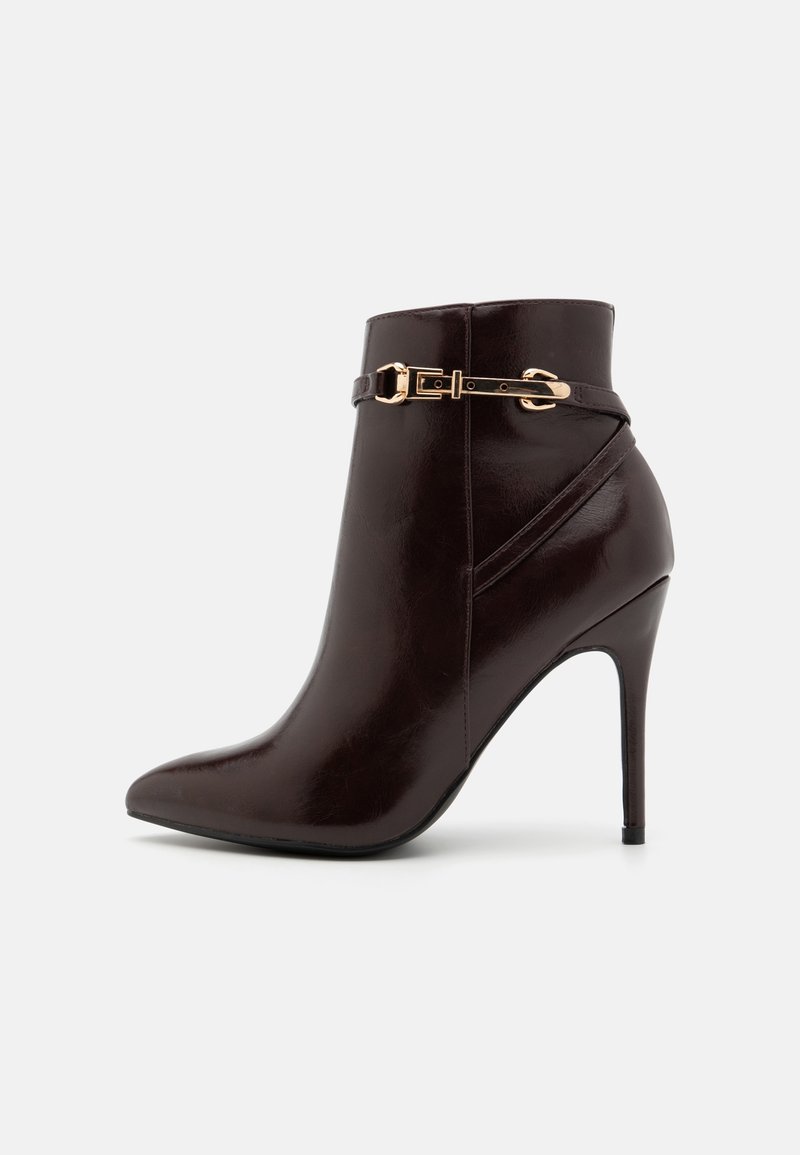 Bottine en cuir marron foncé avec bout pointu, talon aiguille et bride décorative avec boucle dorée autour de la cheville supérieure.