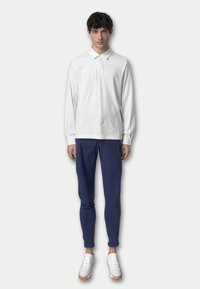 Polo bianco a maniche lunghe con colletto, abbinato a pantaloni blu navy e scarpe bianche. Il polo presenta un piccolo logo sul petto.