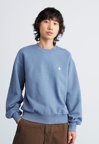Person with curly hair wearing a blue long-sleeve sweatshirt and brown pants, standing against a light background.