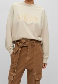 Beige-colourinen collegepaita, jossa "LONDON" on painettu vaaleanruskealla, yhdistetty ruskeisiin samettihousuihin, joissa on vyötärövyö ja useita taskuja.