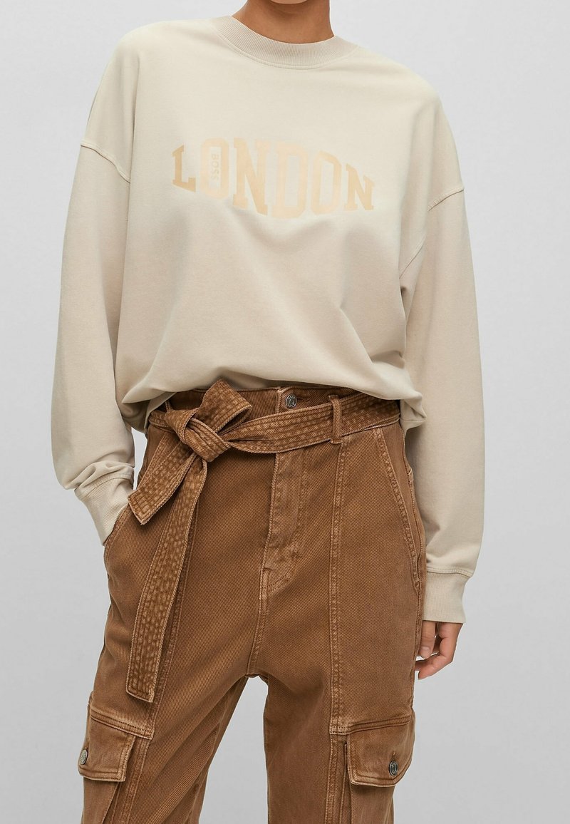 Beige-colourinen collegepaita, jossa "LONDON" on painettu vaaleanruskealla, yhdistetty ruskeisiin samettihousuihin, joissa on vyötärövyö ja useita taskuja.