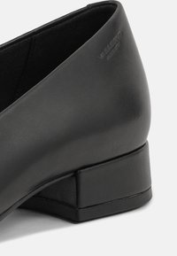 Chaussure en cuir noir à talon bas avec une texture lisse. Le talon est de forme carrée et solide, avec un logo discret sur le côté.