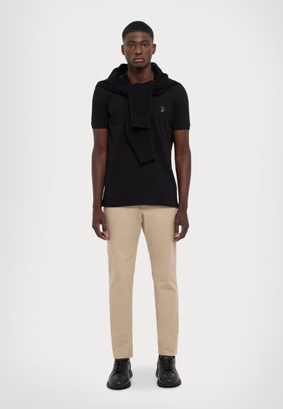Polo preto de mangas curtas, suéter preto de malha drapeado sobre os ombros, calças bege e sapatilhas pretas. Design limpo e moderno.