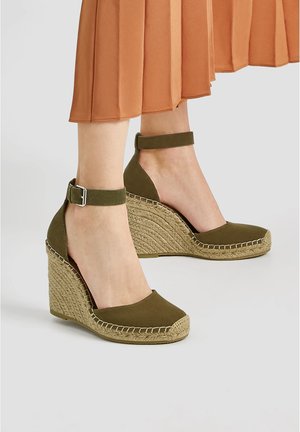 Sandales compensées espadrilles vert olive avec brides à la cheville portées avec une jupe plissée orange rouille.