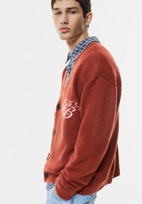 Kardigan in Rostfarbe aus strukturiertem Stoff, mit V-Ausschnitt, schwarzen Knöpfen und einem weißen, gestickten Muster auf der Vorderseite.