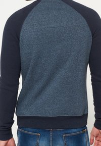 Mörkblå sweatshirt med en strukturerad blå kropp, kontrasterande mörkblå raglanärmar och ribbad fåll. Inga synliga logotyper eller mönster.