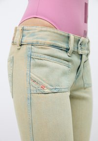Jeans bleu clair délavé avec une étiquette Diesel Industry, porté avec un haut rose ajusté, montré de profil au niveau de la taille et des hanches.