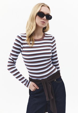 Mujer rubia con gafas de sol ovaladas, una camisa de rayas largas en marrón, blanco y azul, jeans oscuros y un suéter oscuro atado a la cintura.