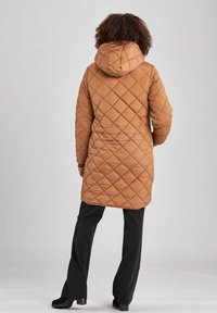 Parka iz prešitih rjavih materialov s kapuco, ki se ponaša z dolgim silhuetom in diagonalnim šivom. V kombinaciji s črnimi hlačami in elegantnimi čevlji.
