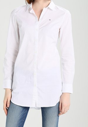 Femme portant une longue chemise blanche boutonnée avec col et un jean bleu, debout devant un fond clair uni.