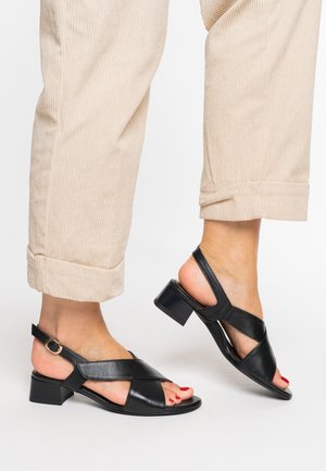 Schwarze Slingback-Sandalen aus Leder mit überkreuzten Riemen, niedrigem Blockabsatz und einem goldenen Schnallendetail, kombiniert mit hochgekrempelten beigen Cordhosen.