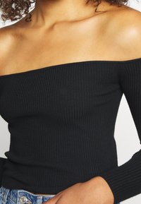 Svart off-shoulder ribbad topp med långa ärmar, med en tät stickad struktur och figurnära silhuett. Tyget ser mjukt och elastiskt ut.