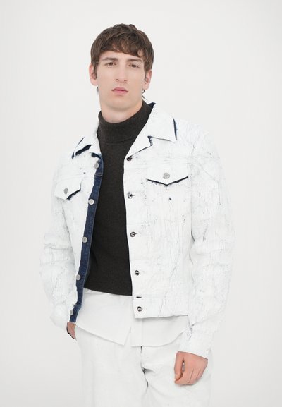 White denim jacket with textured finish, front pockets, and dark blue accents. Worn over a dark turtleneck and layered white shirt.