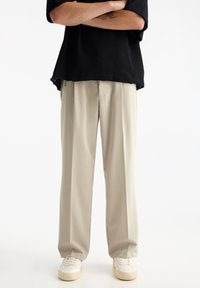 Beige Weite-Leg-Hosen mit glatter Textur und hohem Taillenbund, kombiniert mit einem lockeren schwarzen Strickoberteil. Weiße Sneakers runden das Outfit ab.