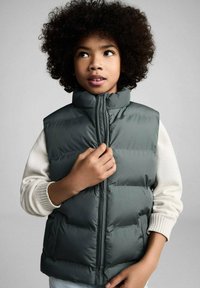 Gilet verde imbottito con colletto alto, chiusura con zip e motivo a righe orizzontali. Indossato sopra un maglione leggero con polsini a coste.