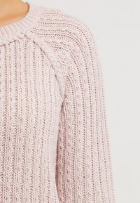 Ljusrosa stickad tröja med ribbad struktur och flätad detalj längs axelsömmen. Tyget verkar mjukt och elastiskt.