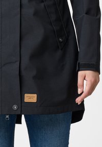 Black waterproof jacket with a zip closure, snap buttons, adjustable cuffs, and a branded patch on the front. Features smooth fabric texture.