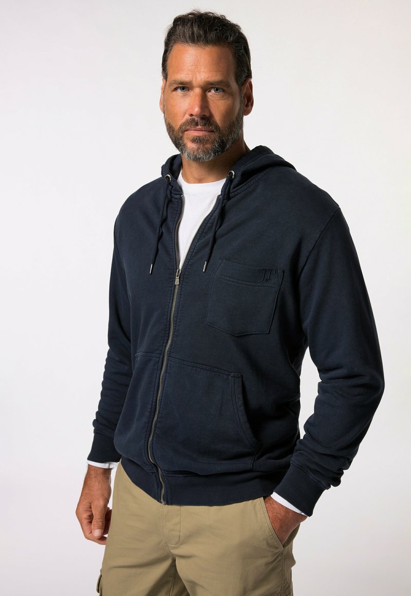 Navy blue zip-up hoodie, featuring a front pocket, drawstring hood, and ribbed cuffs and hem. Fabric appears textured and soft.