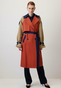 Abrigo de trench de color naranja con acentos azules y mangas beige. Presenta un frente de doble botonadura, cintura ceñida con cinturón y un gran cuello.
