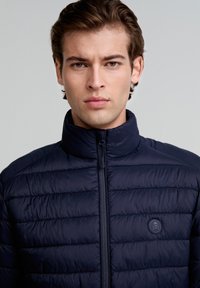 Chaqueta de abrigo de color marino con cuello doblado, textura acolchada horizontal, cremallera completa y un parche con un logo que presenta una calavera y huesos cruzados en el pecho.