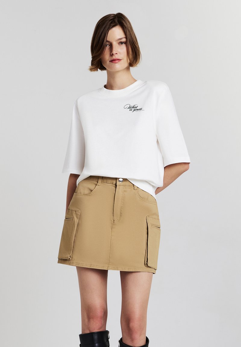 T-shirt en coton blanc à manches courtes avec texte noir. Jupe beige avec deux poches latérales, taille haute et texture lisse. Bottes noires arrivant au genou.