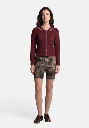 Frau trägt eine burgunderrote Strickjacke mit Knöpfen, traditionelle bestickte braune Lederhose und dunkelbraune Oxford-Schuhe, steht aufrecht.