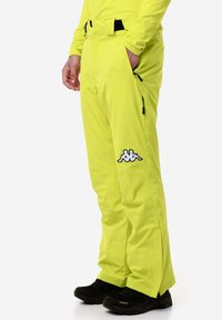 Pantaloni da sci gialli brillanti con vita regolabile, tasche con zip, cuciture rinforzate e un logo bianco sulla coscia sinistra.