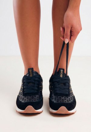 Persona atando zapatillas negras con recortes estampados, de pie sobre una superficie neutra, con los cordones en la mano derecha.