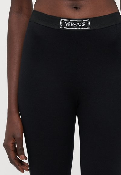 Colanți negri cu un brâu elastic lat, având un eticheta albă cu logo-ul "VERSACE". Textură netedă, design mulat.