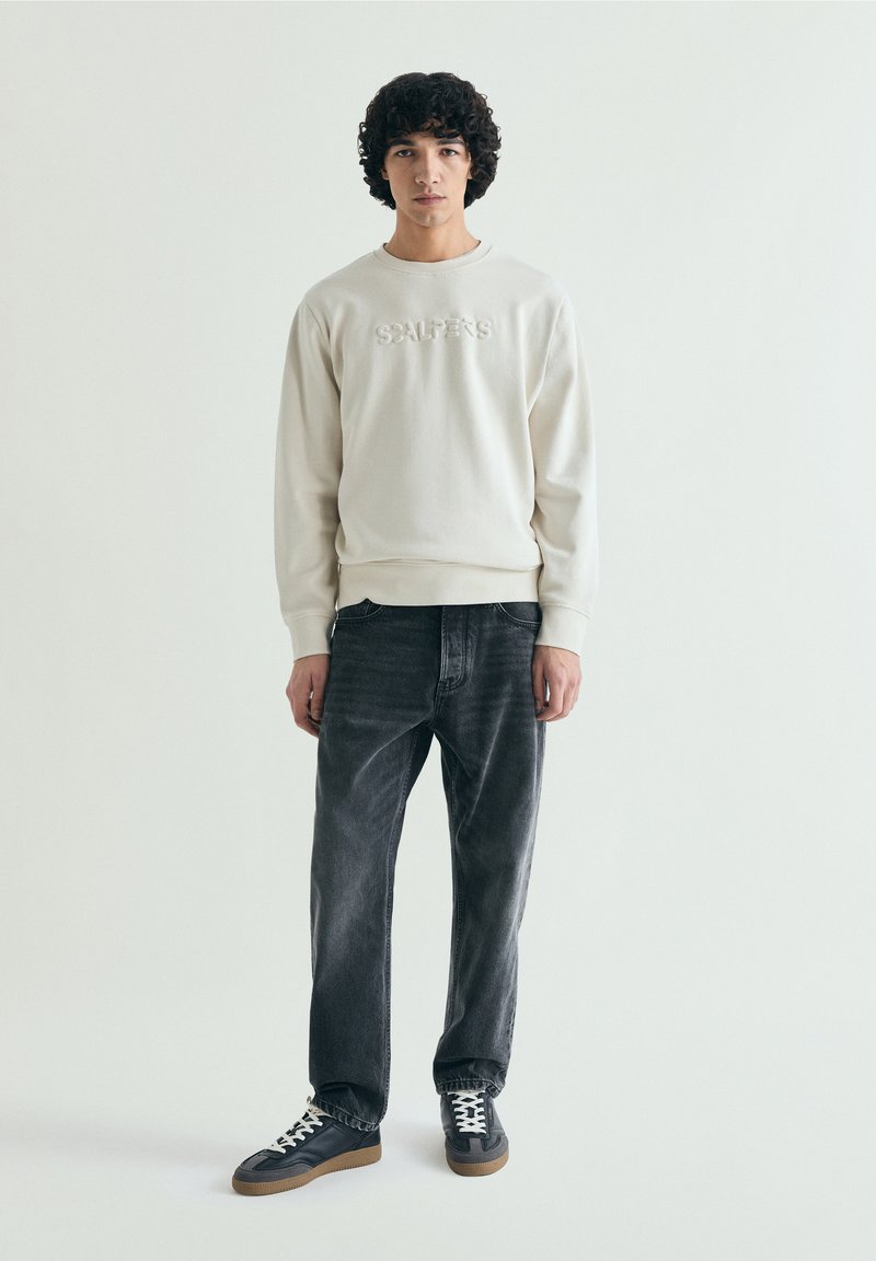 Jeune homme aux cheveux bouclés portant un sweat-shirt écru avec un texte embossé, un jean gris foncé et des baskets noires à lacets blancs.