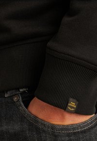 Sudadera negra con puños acanalados, que presenta una pequeña etiqueta de logo amarilla. Combinada con unos jeans de denim oscuros, mostrando un contraste de texturas.