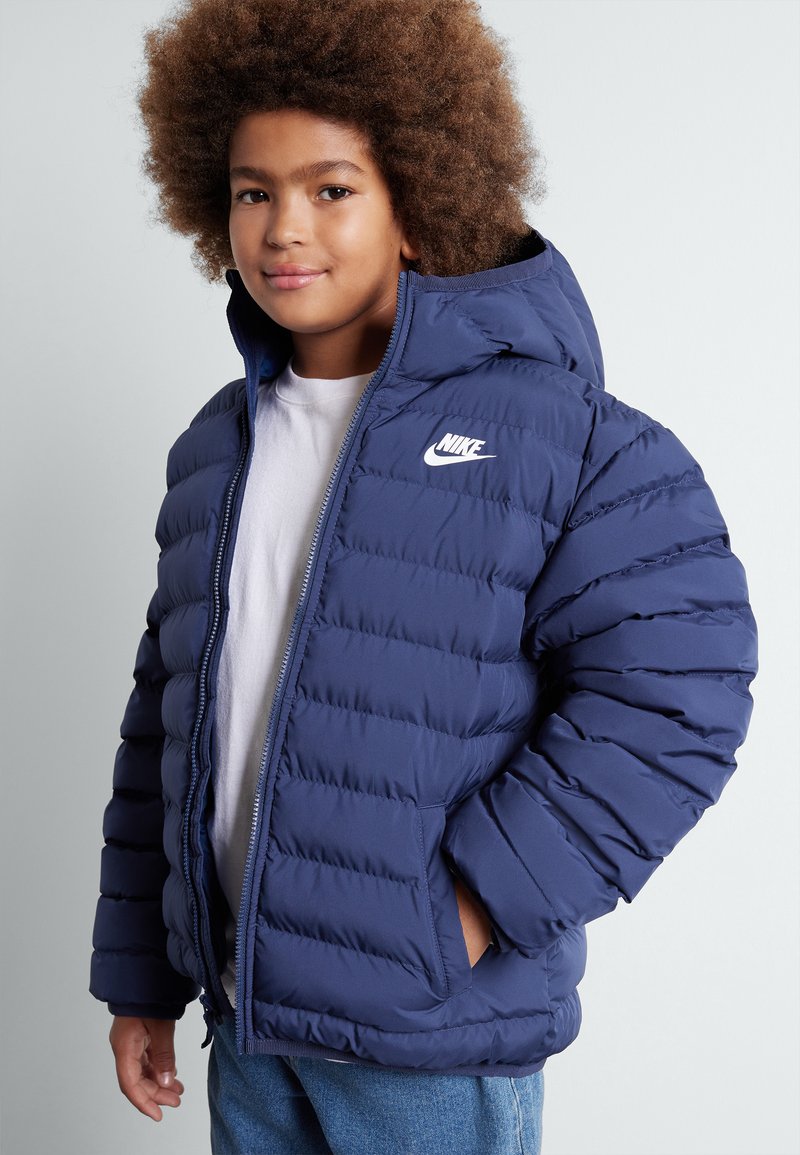 Giacca puffer blu navy con cappuccio, chiusura con zip e logo Nike sul petto. Presenta una texture trapuntata e tasche laterali.