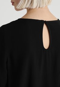 Blond kvinna sedd bakifrån som bär en svart topp med en liten tårformad nyckelhålöppning som är fastsatt med en knapp på övre sidan av ryggen.