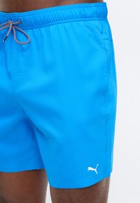 Ανδρικό μπλε φωτεινό μαγιό με μαύρο και λευκό κορδόνι και λευκό λογότυπο Puma στο αριστερό πόδι, φορεμένο από άτομο με σκούρο δέρμα.