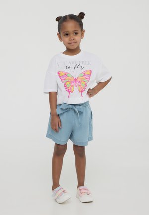 T-shirt bianco a maniche corte con grafica di farfalla rosa e arancione, abbinato a pantaloni denim blu con dettagli a fiocco. Sneakers rosa.