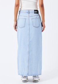Falda maxi de mezclilla azul claro con corte recto, dos bolsillos traseros y una costura cosida que corre por el centro. Llevada con zapatillas blancas.