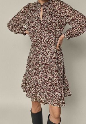 Kobieta ubrana w sukienkę z długim rękawem, sięgającą do kolan, w brązowy kwiatowy wzór z falbanami na dole, do tego czarne kozaki za kolano.