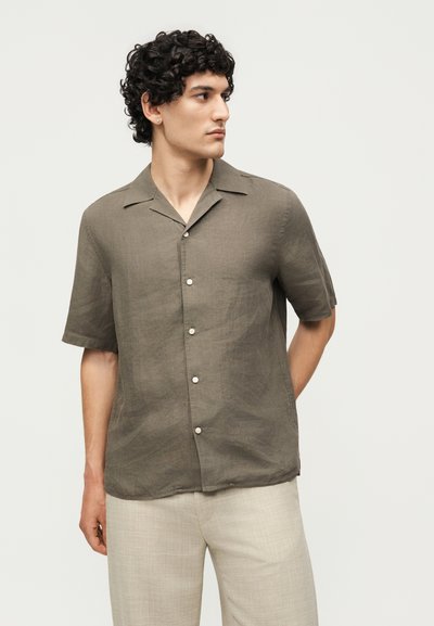 Άνδρας με σγουρά μαλλιά που φοράει μία ελιάς κοντομάνικη πουκάμισα με κουμπιά και ανοιχτό μπεζ παντελόνι, κοιτάζοντας αριστερά με το ένα χέρι πίσω από την πλάτη.