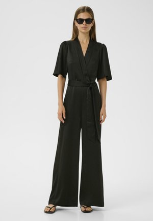 Schwarzer Jumpsuit mit kurzen Ärmeln, Wickeldesign, weitem Bein und einem gebundenen Taillengürtel. Glatte Textur, kombiniert mit schwarzen Sonnenbrillen und Flip-Flops.